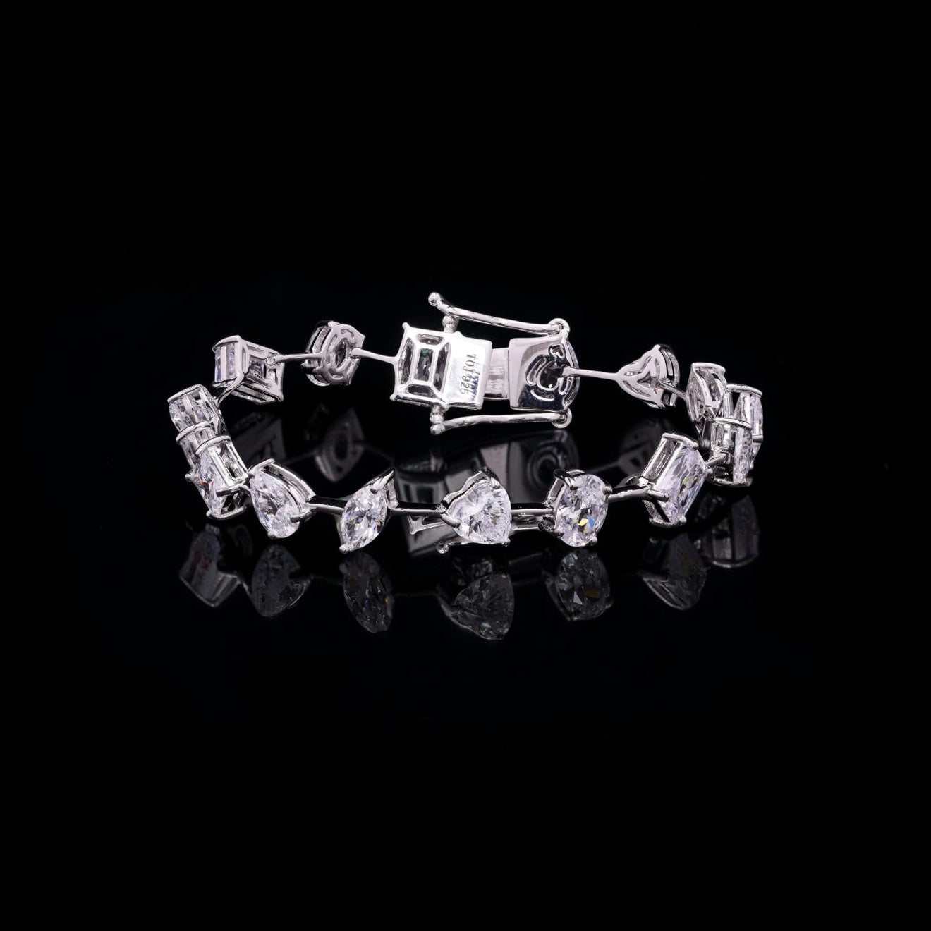 A silver tennis bracelet featuring a mix of round, baguette, and marquise-cut clear stones, displayed on a reflective black surface against a solid black background.