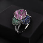 A silver-toned ring with three overlapping flower-like faces set with pink, green, and blue pavé stones, displayed on a black surface.