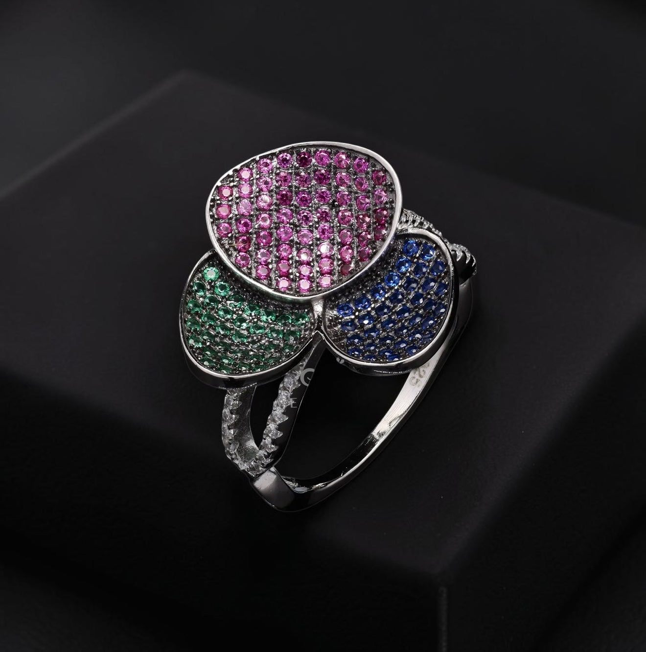 A silver-toned ring with three overlapping flower-like faces set with pink, green, and blue pavé stones, displayed on a black surface.